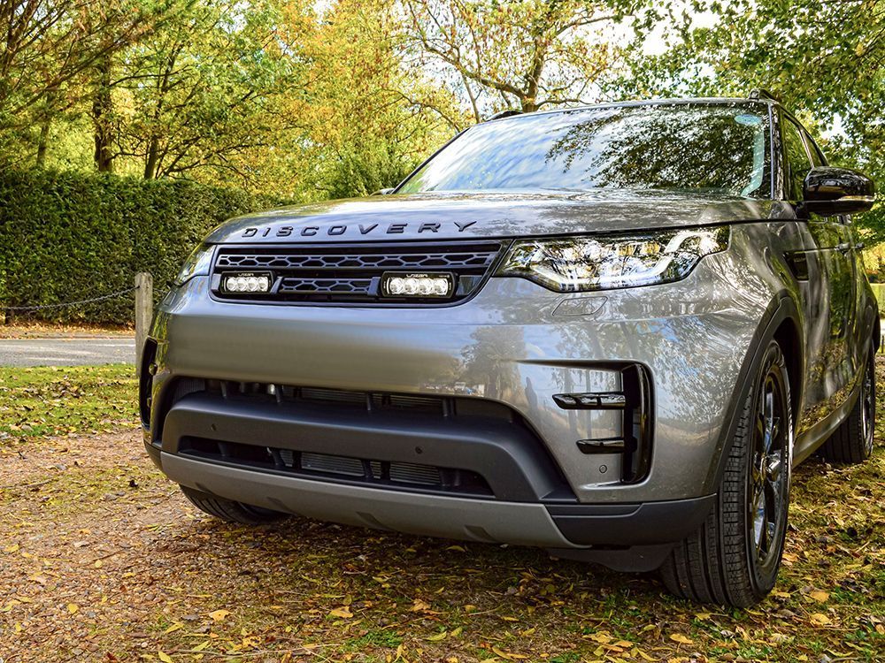 Silver Land Rover Discovery parked on a dirt path, facing the camera with a green hedge and trees in the background.