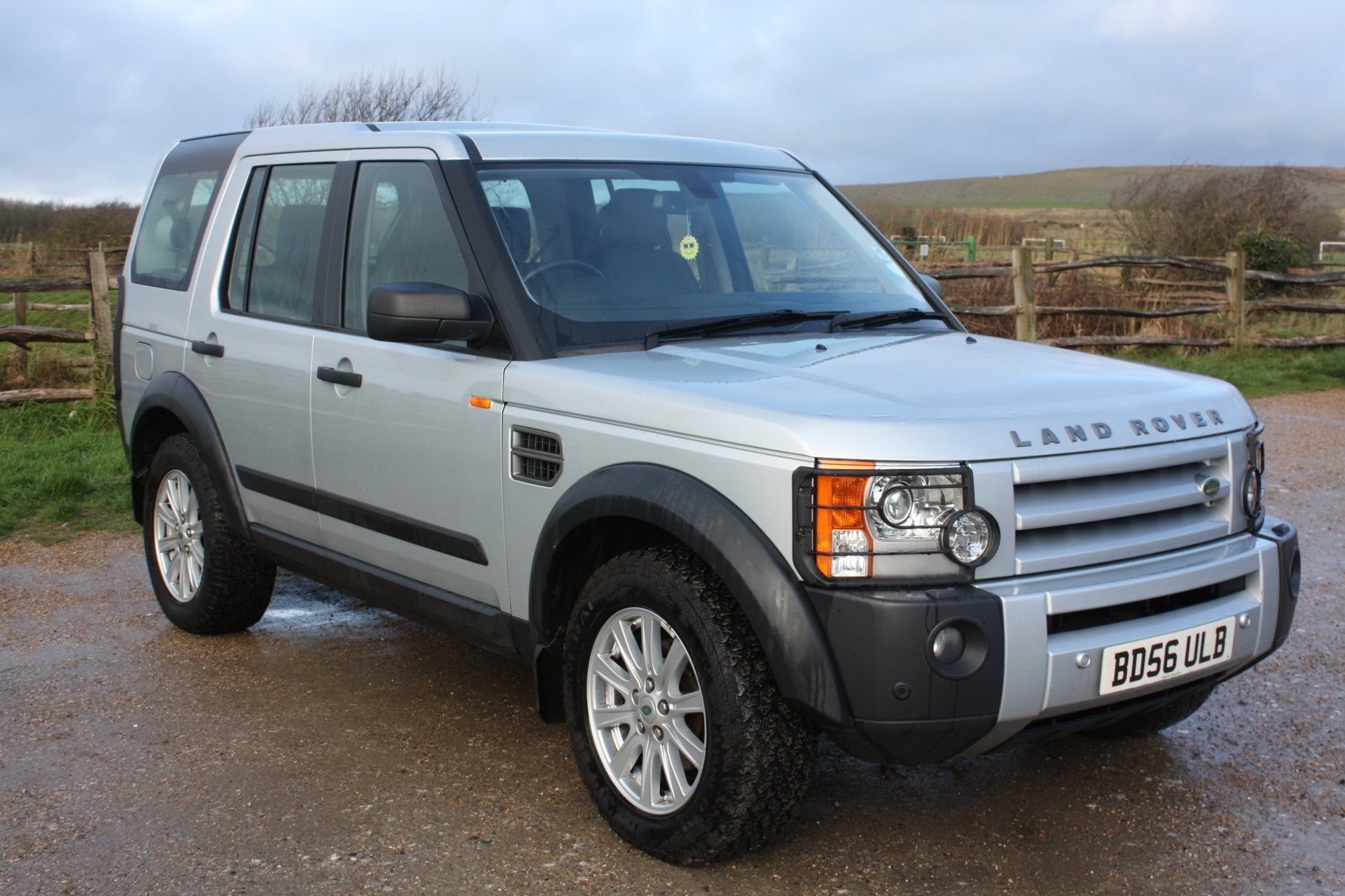 Silver Land Rover Discovery SUV parked on a paved surface in front of a wooden fence and field under a cloudy sky.