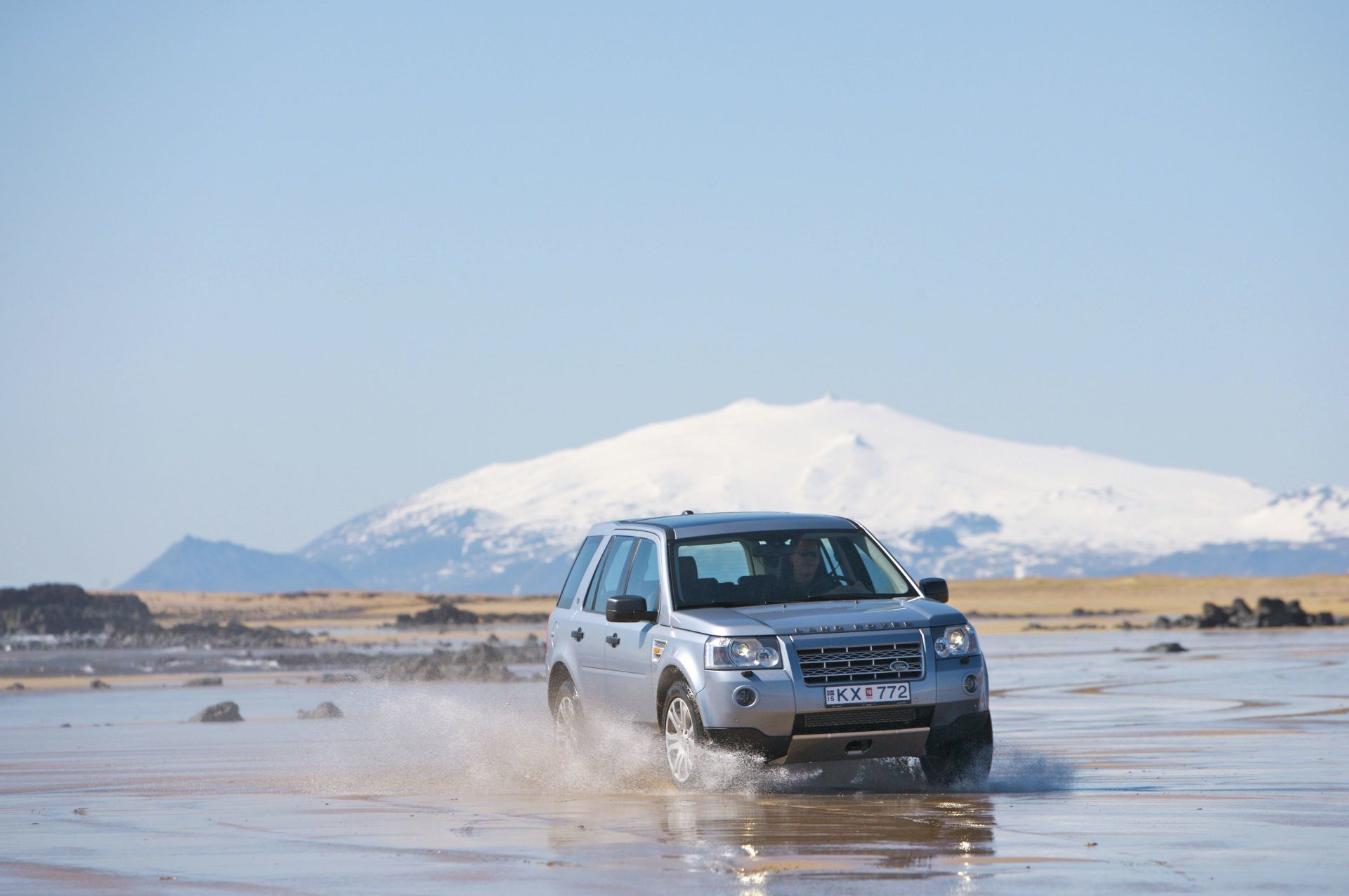 Silver SUV fording shallow water with a snow-capped mountain in the background under a clear blue sky.