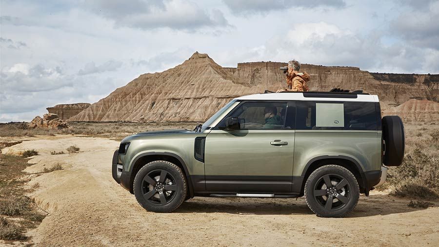 Green Land Rover Defender parked in a desert landscape. A person stands on the roof, overlooking the scenery.