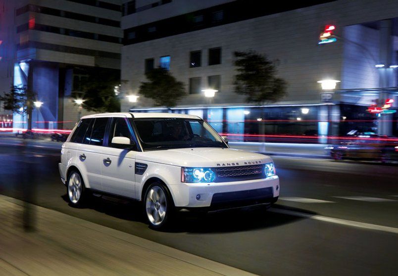 White Range Rover Sport driving on a city street at night. Headlights and streetlights illuminate the scene.
