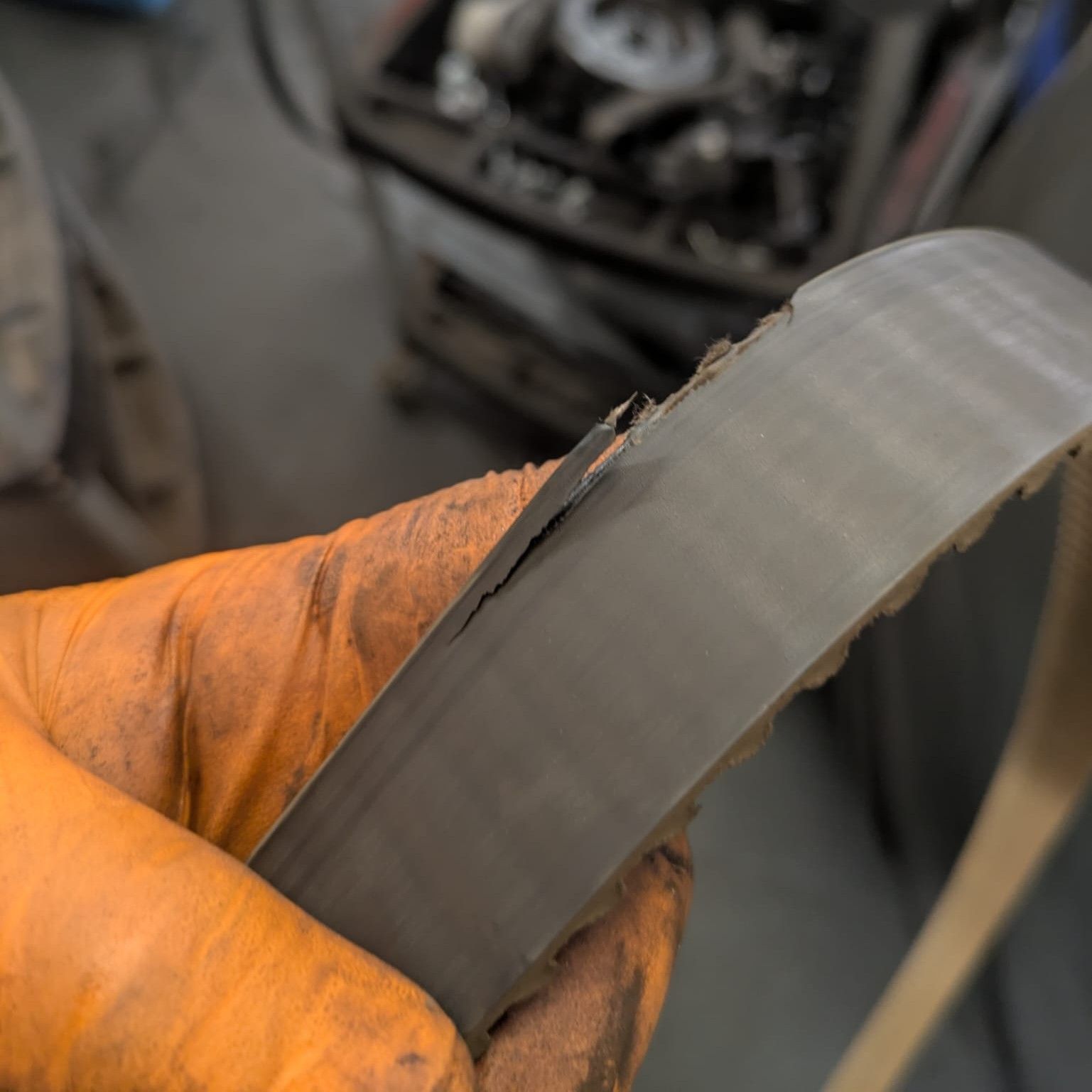 A gloved hand holds a worn, cracked timing belt, likely from a car engine, in a garage setting.