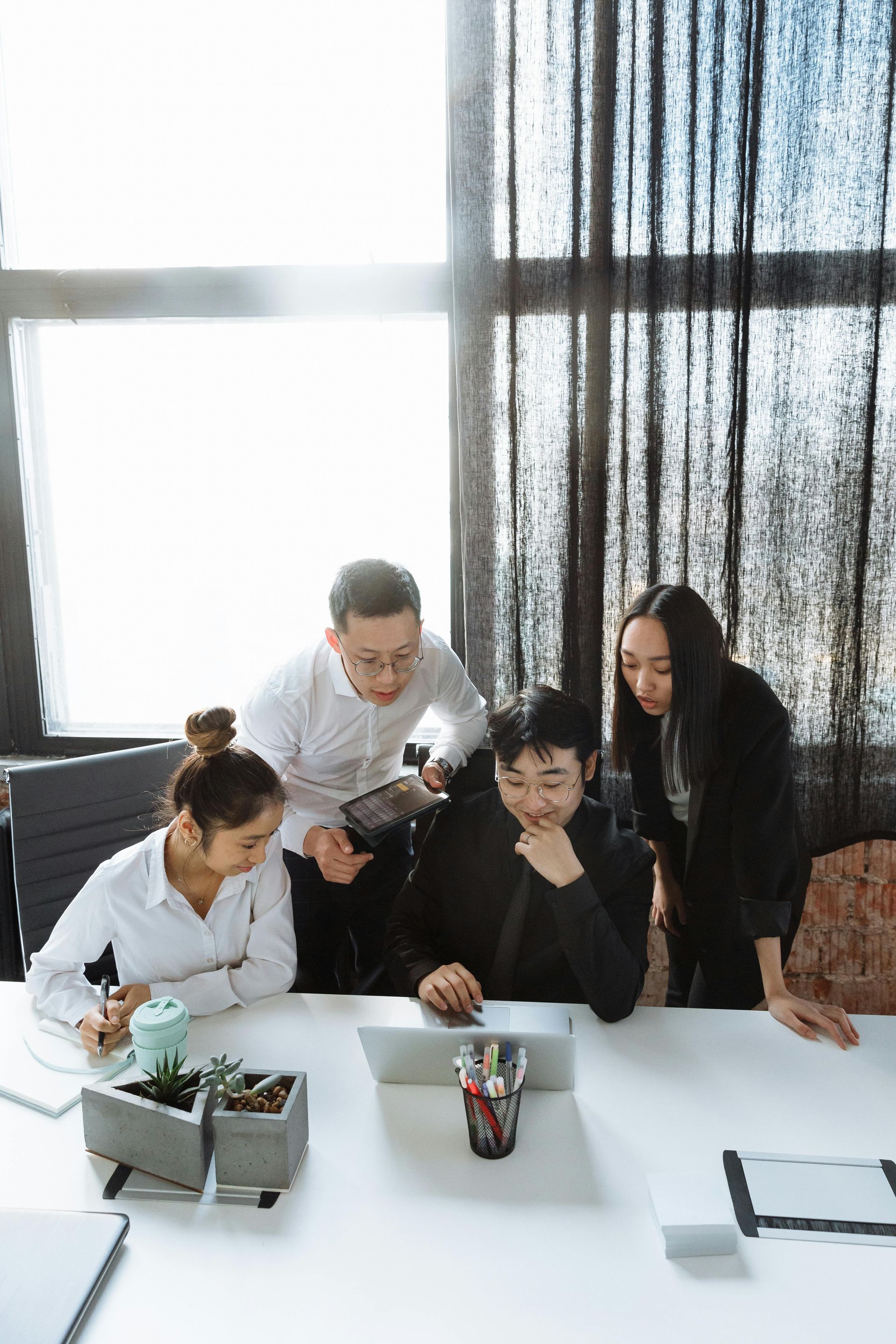 Four coworkers gather around a laptop at a bright office desk, reviewing documents and smiling.