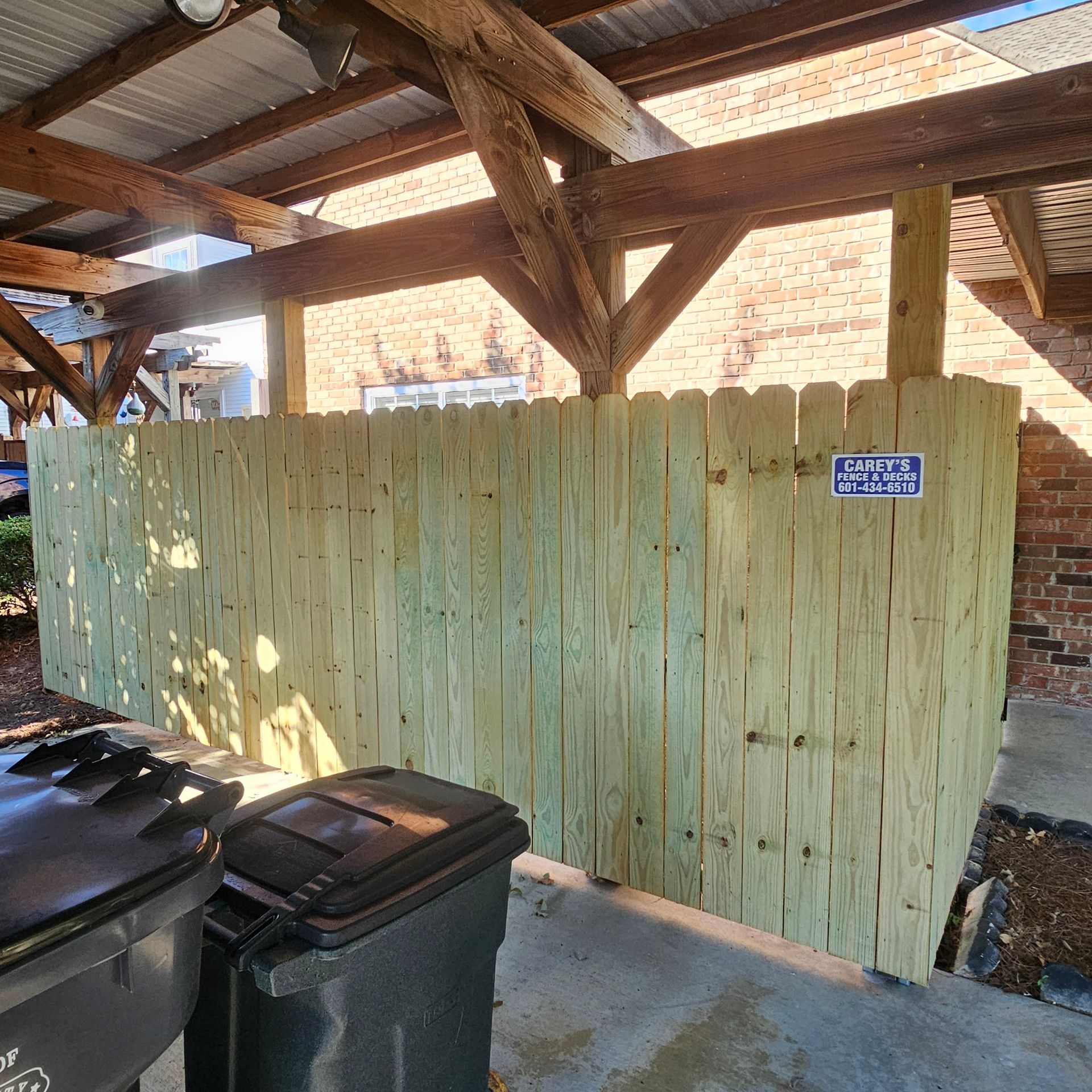 A wooden fence is surrounded by trash cans and a brick building.