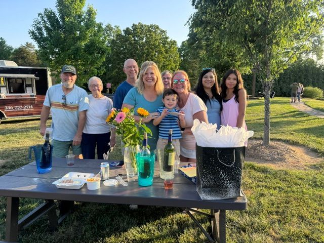 Group of people smiling outdoors by a picnic table with drinks and flowers in a sunny park