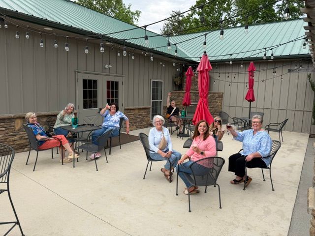 People seated around tables on a patio outside a building, with red umbrellas and a metal roof.