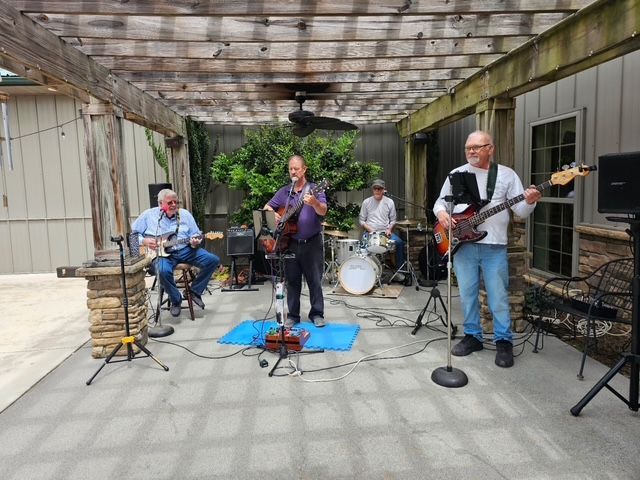Four musicians performing under a patio canopy with drums, guitars, and microphones
