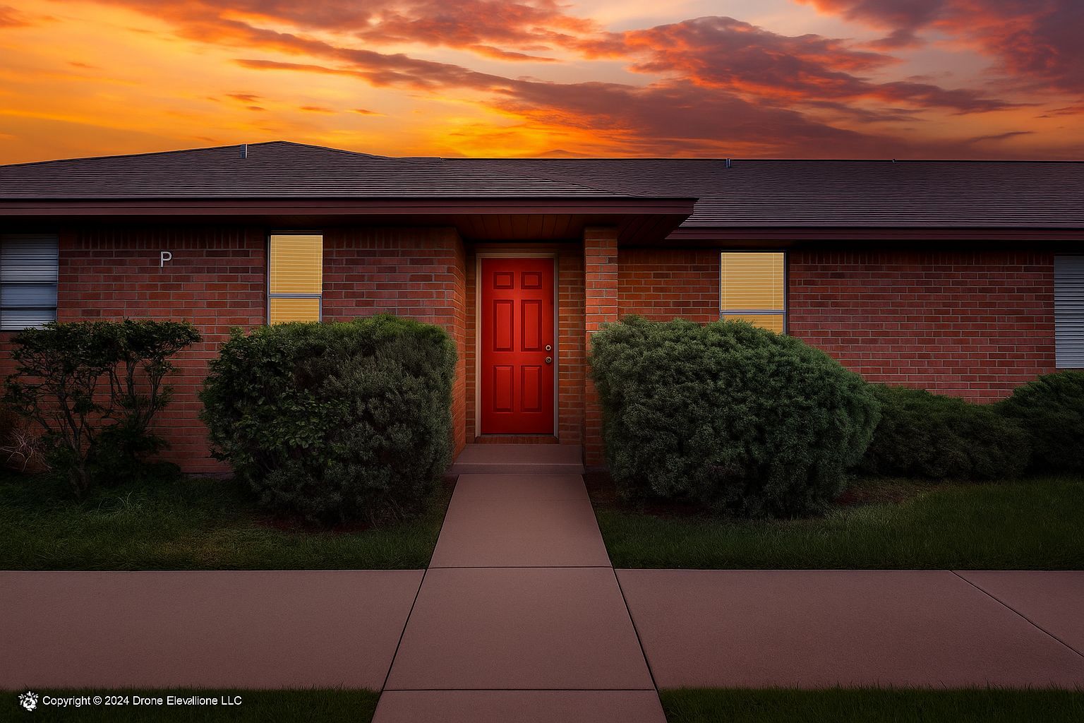 Red door on a brick house, flanked by green bushes, under an orange sunset.