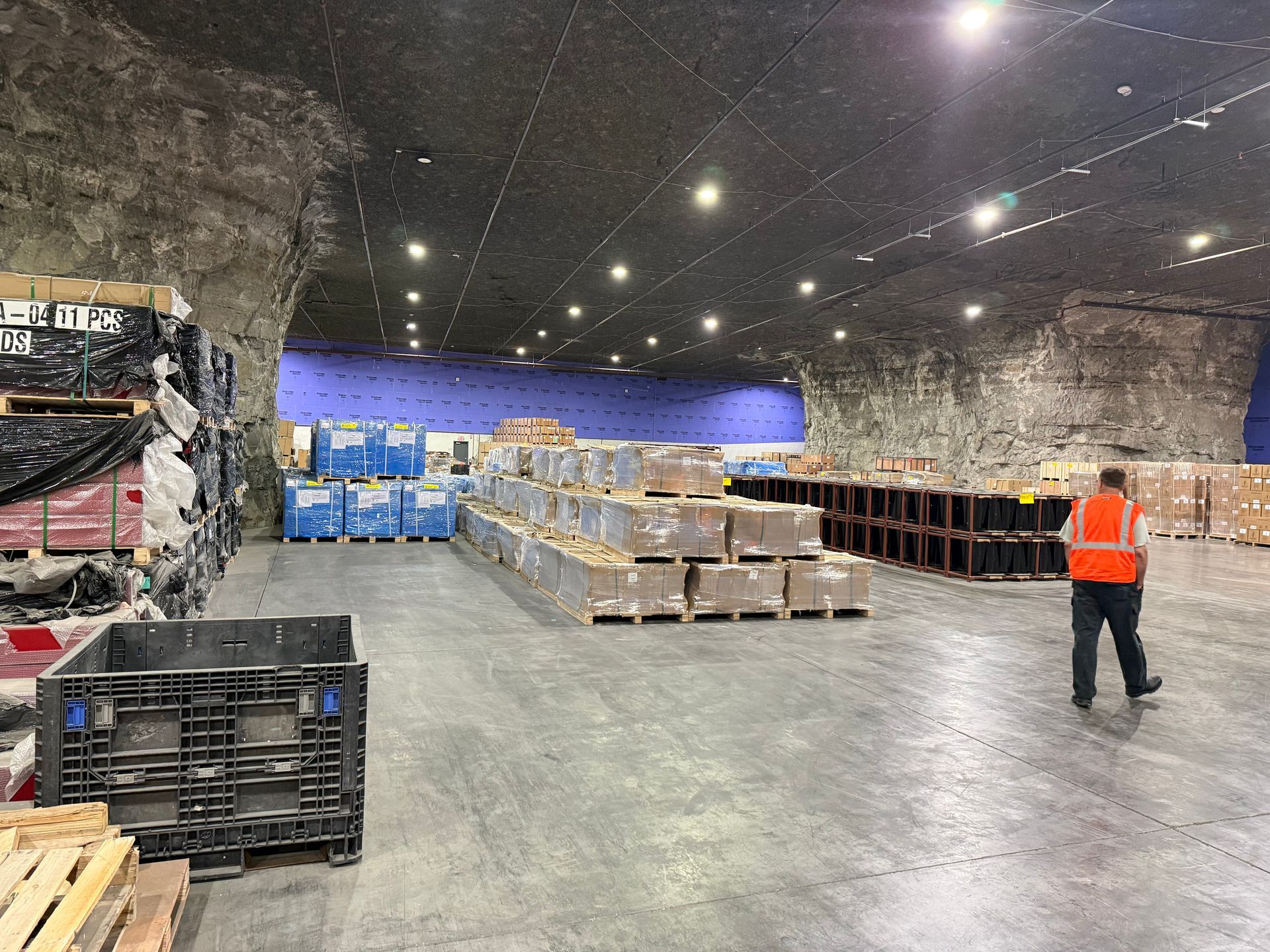 Warehouse interior with pallets and crates, blue-lit back wall, and a worker in an orange safety vest walking.