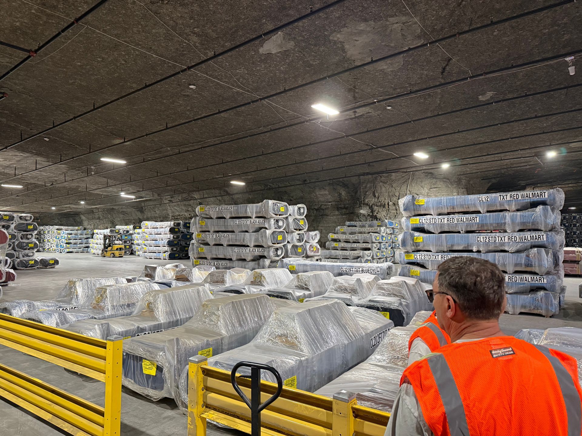 Warehouse with stacked wrapped pallets; worker in orange safety vest looks over the storage area.