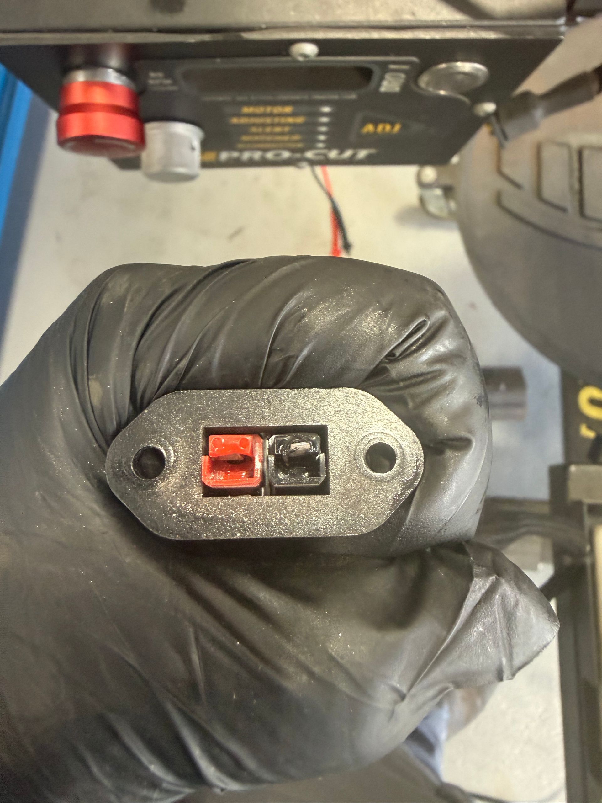 Gloved hand holding a black two-pin electrical connector with red and black terminals near a device assembly