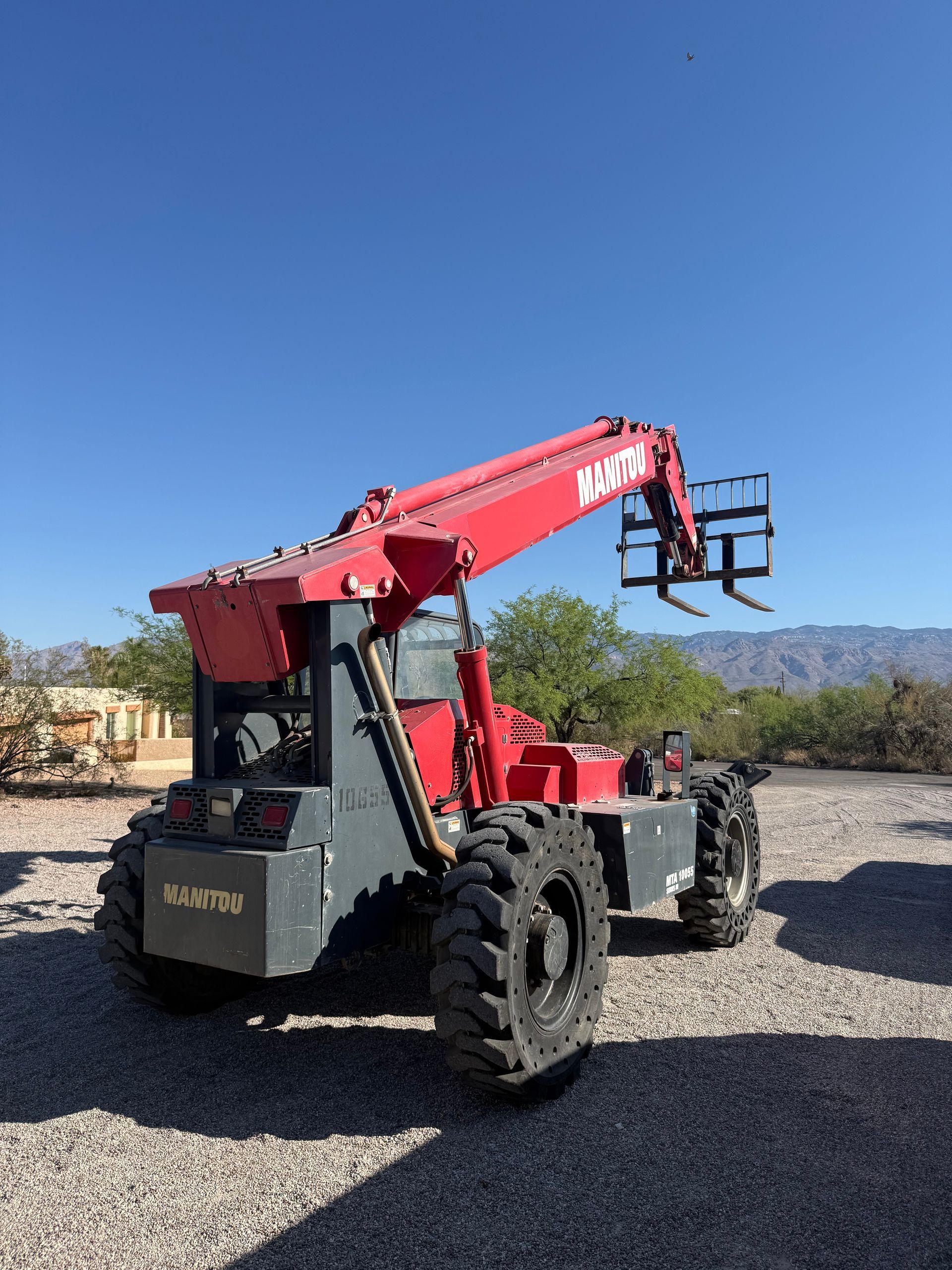 Red telehandler forklift parked on gravel under a clear blue sky, with mountains in the background
