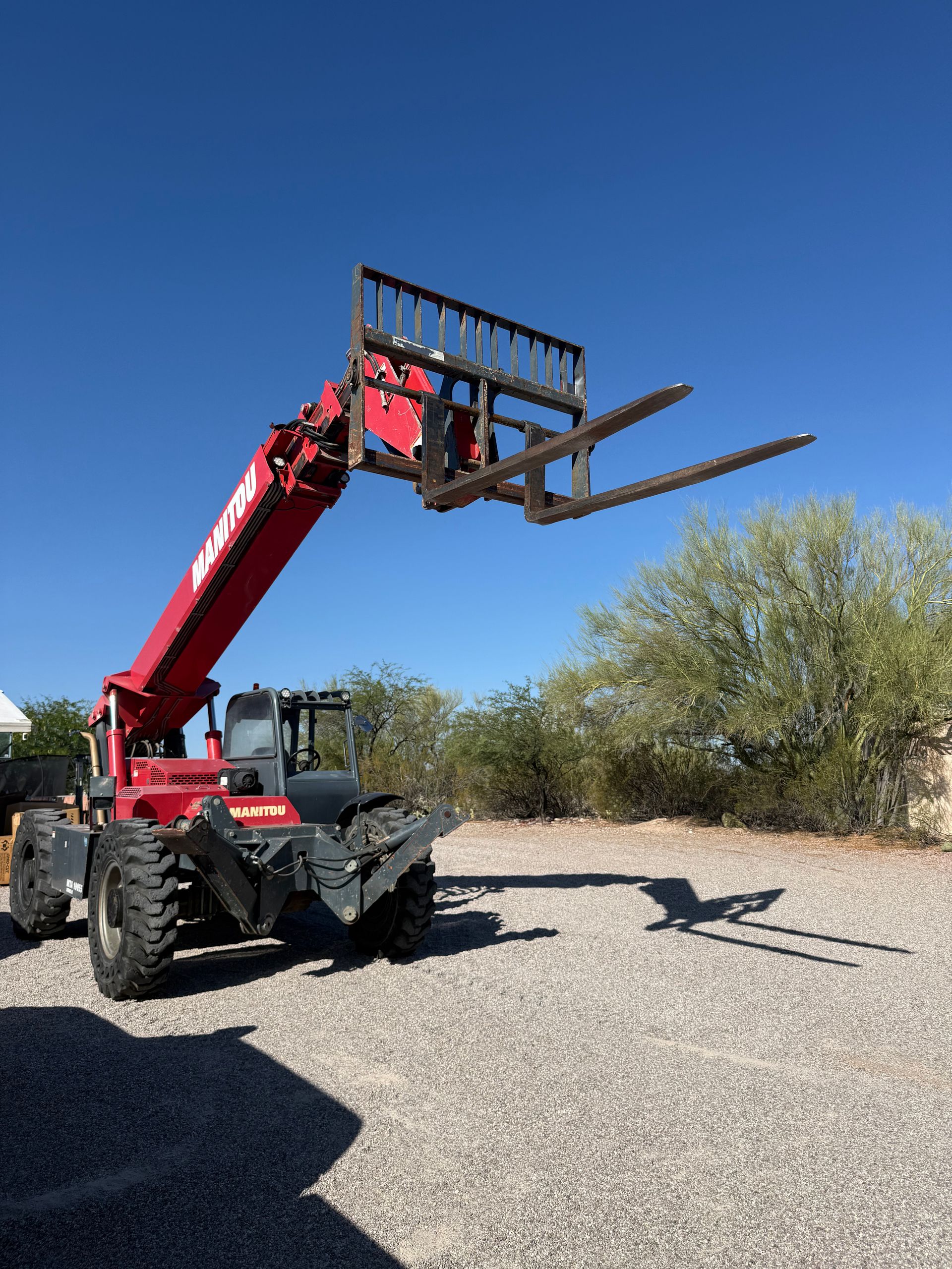 Red telescopic forklift with raised pallet forks on gravel under a clear blue sky.