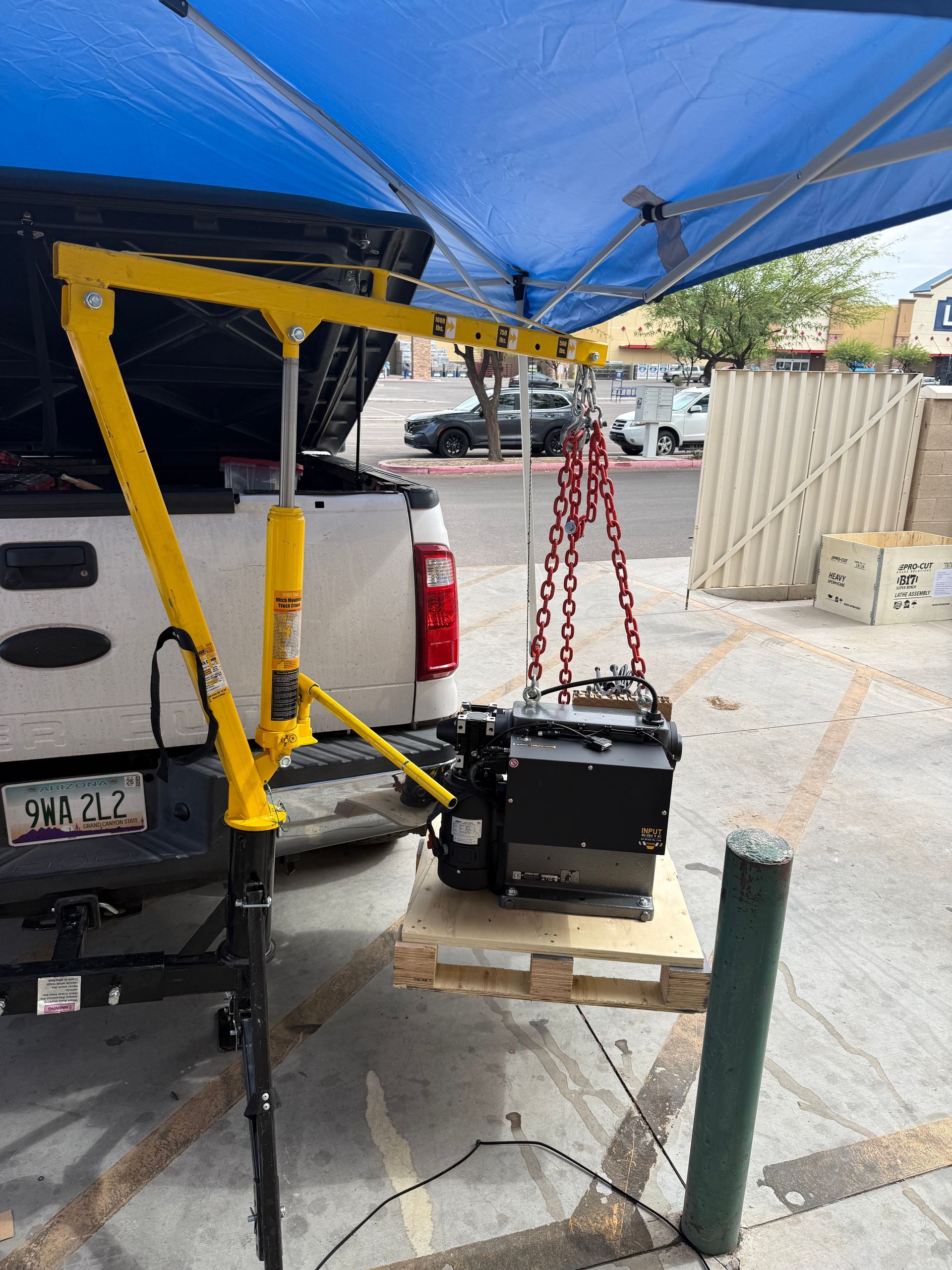 Portable lift hoists a black box from a pickup truck under a blue canopy.