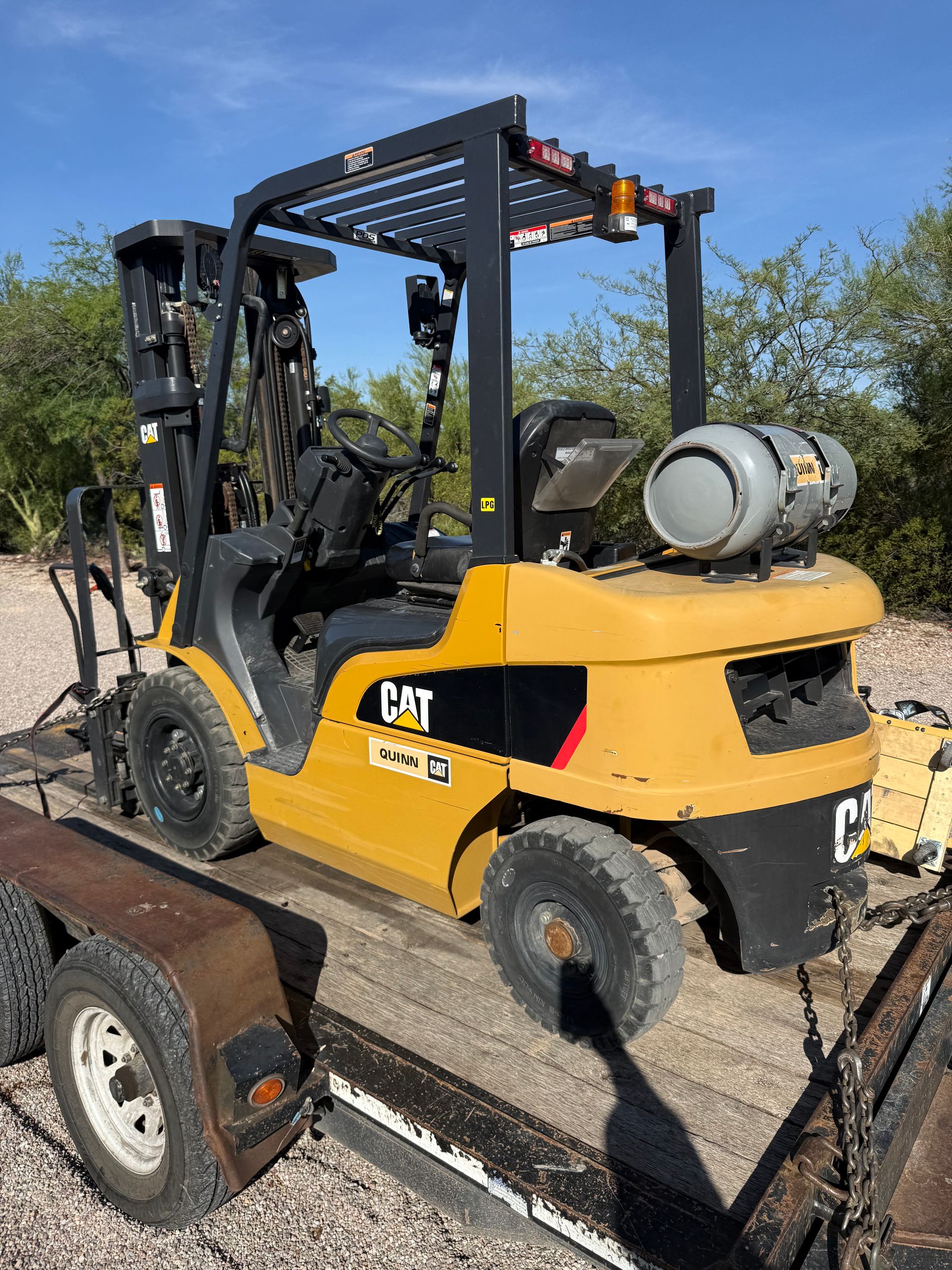Yellow CAT forklift on a trailer outdoors, carrying a propane tank under a blue sky.