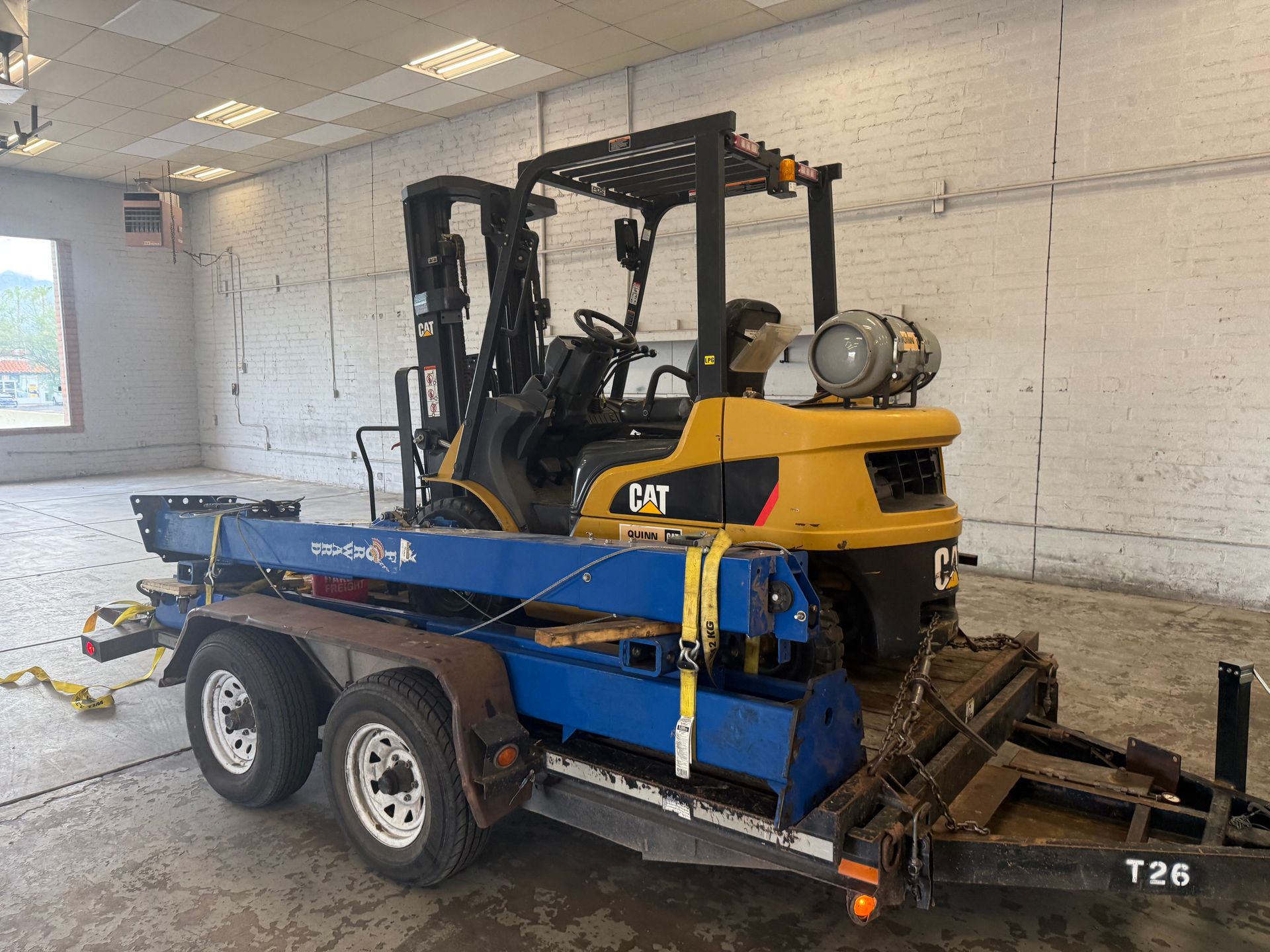 Yellow CAT forklift secured on a blue dual-axle trailer inside a white garage.