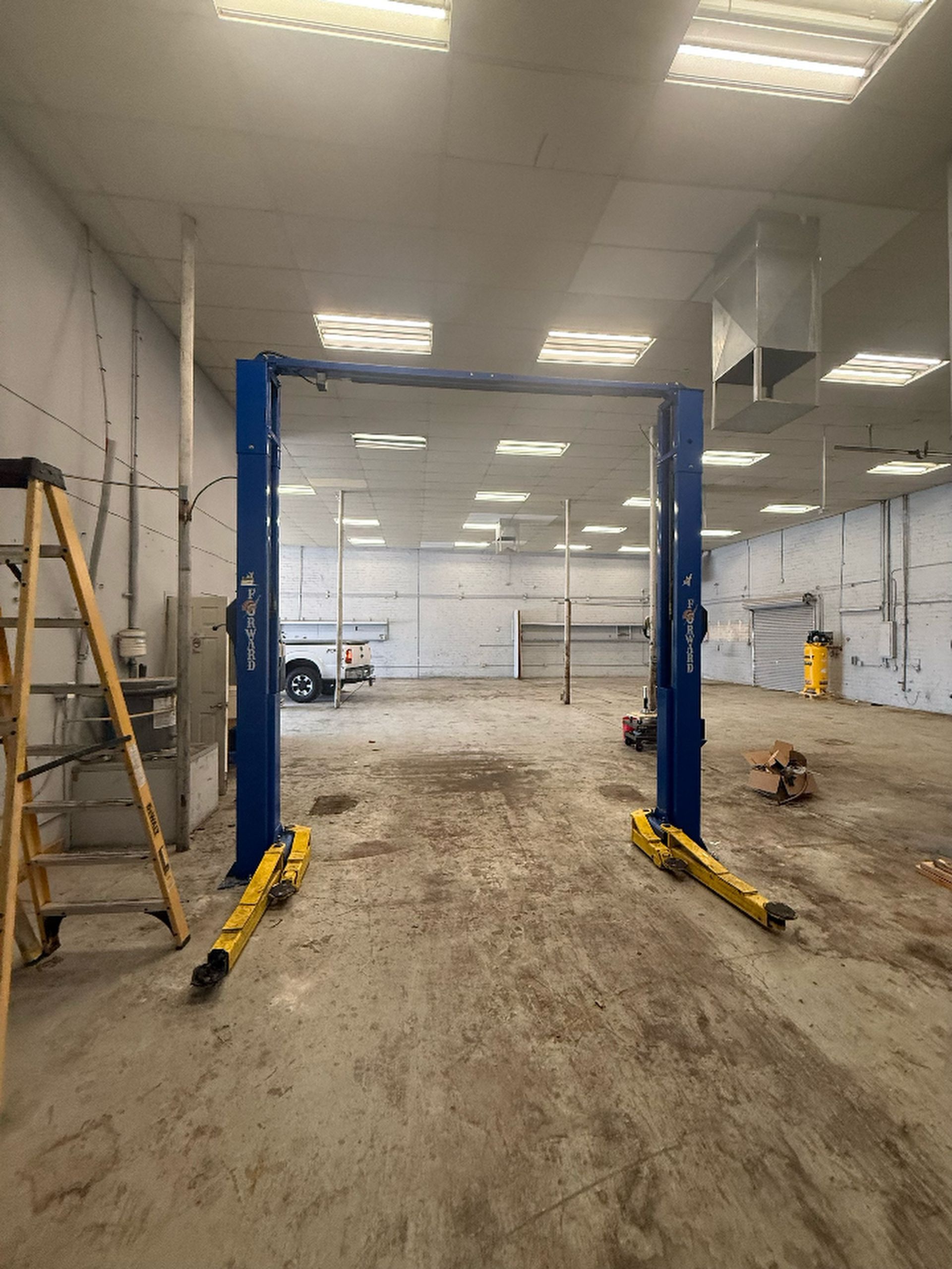 Empty auto shop bay with a blue two-post car lift and a ladder on the left inside a concrete garage.