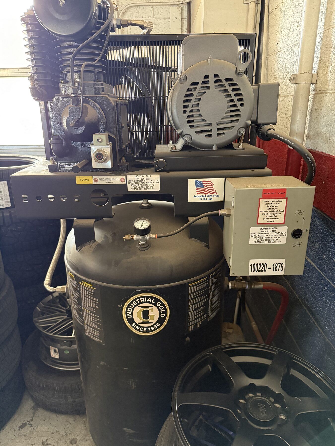 Air compressor with motor and tank in a cluttered garage, with stacked tires and a wheel in front.