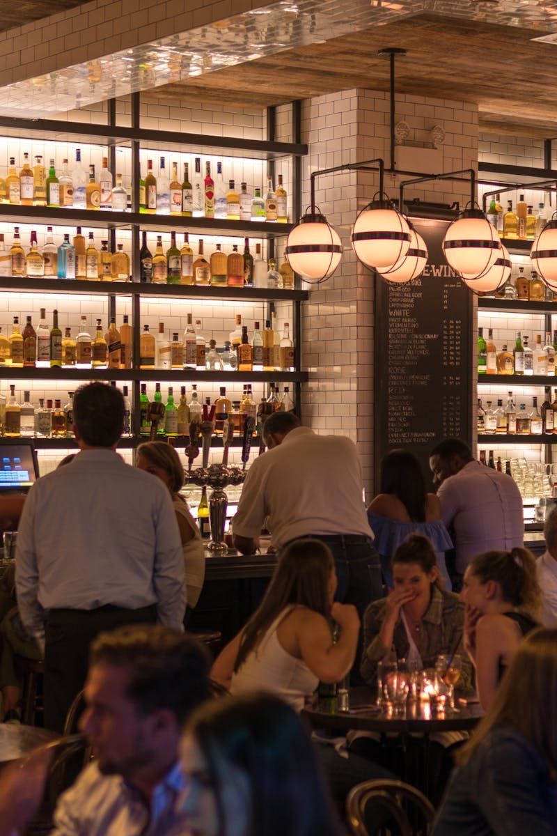 Intérieur de bar avec des clients, des étagères à spiritueux illuminées et des suspensions lumineuses en forme de globe.