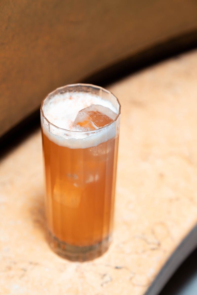 Grand verre de cocktail rouge-orangé avec mousse et glaçons, posé sur un comptoir en marbre.