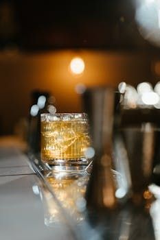Un cocktail sur le comptoir avec des glaçons, des outils de bar flous et un éclairage chaleureux en arrière-plan.