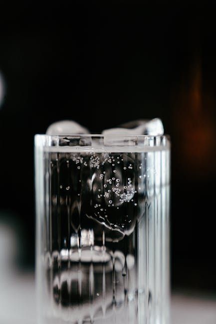 Verre de liquide transparent avec des glaçons et des bulles, dans un verre cannelé, sur fond sombre.