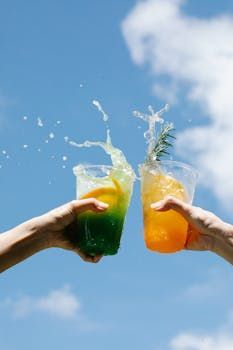 Deux mains font tinter des gobelets en plastique transparent remplis de boissons vertes et orange