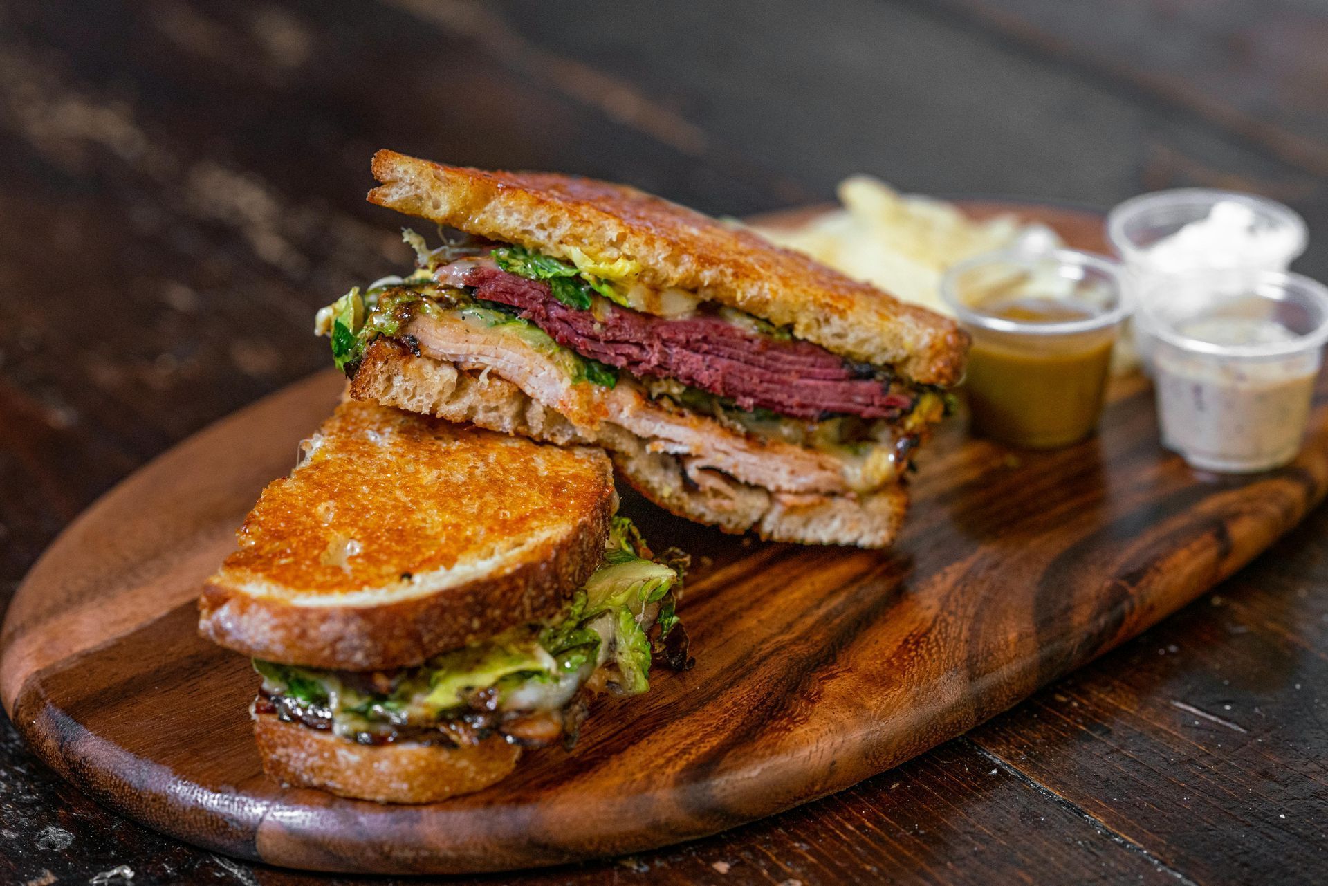 Gourmet burger on a wooden board with dipping sauces, sliced in half to show layers of meat and greens