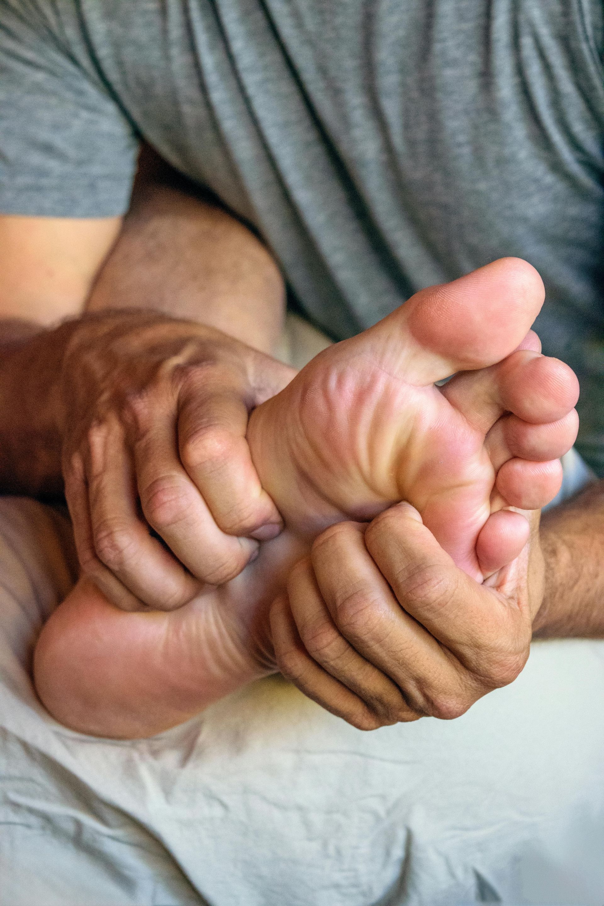 Person holding and massaging a bare foot close-up, with a gray shirt in the background.