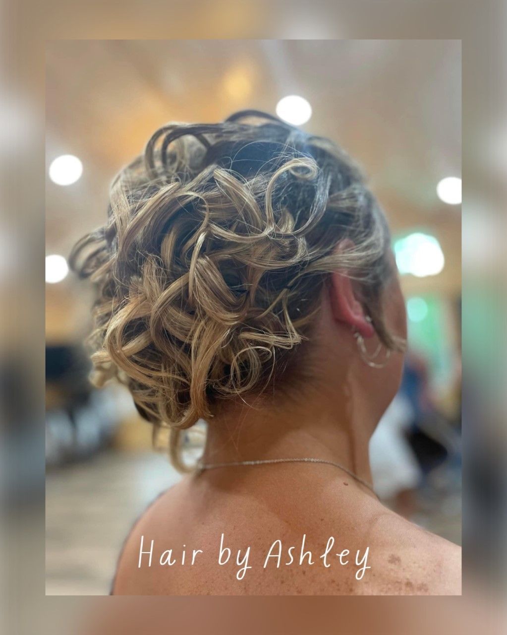 Curly updo hairstyle with blonde highlights and loose tendrils, shown from the back in a salon.