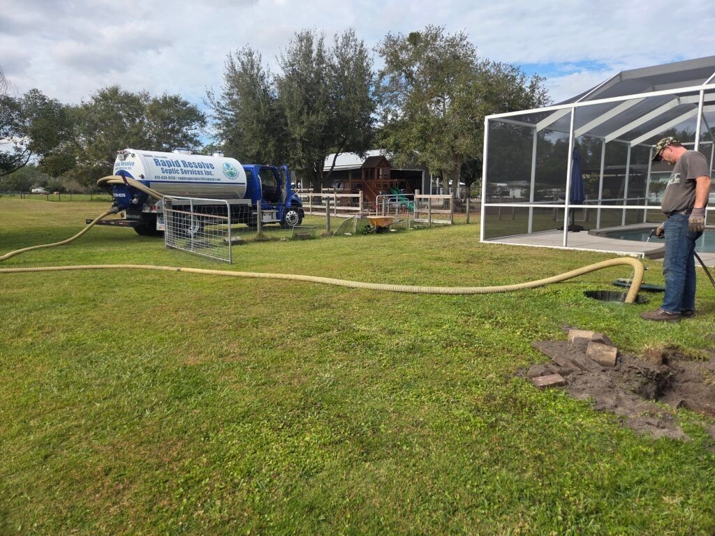 Man watering grass beside a greenhouse while a large blue tank trailer sits in the yard