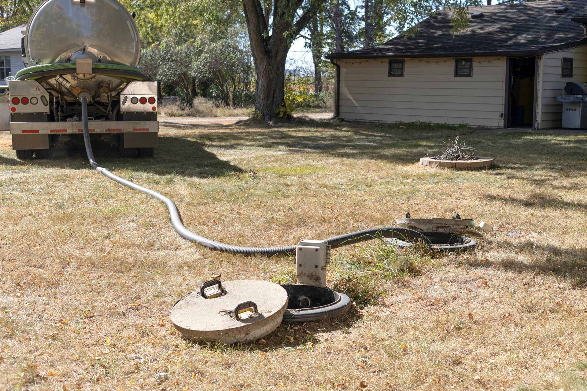 Yard with a hose leading from a tank trailer to an open ground pit beside a small house