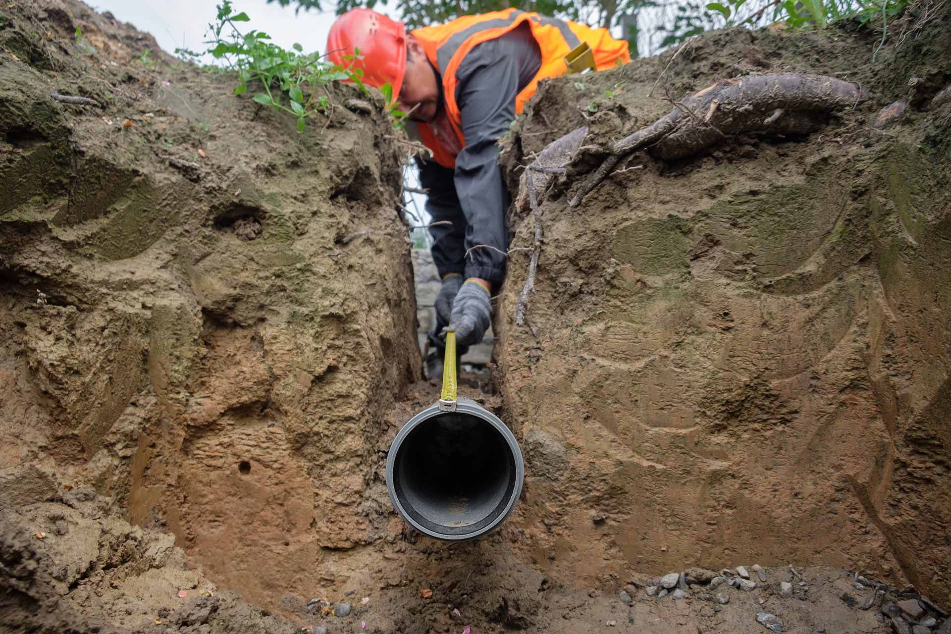 Worker in orange safety gear standing in a trench, holding a large black pipe section.