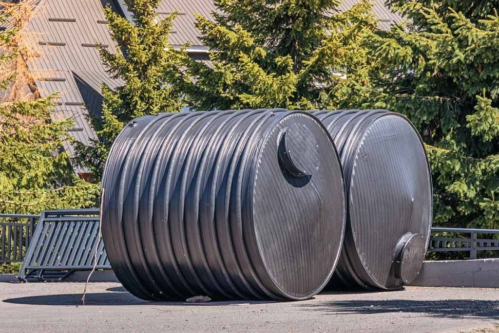 Two large black ribbed cylindrical sculptures outdoors on a paved area near trees and benches