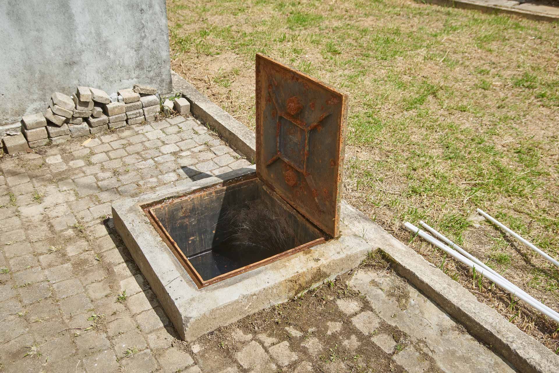 Open metal utility hatch in a cobblestone patio beside grass and a wall.