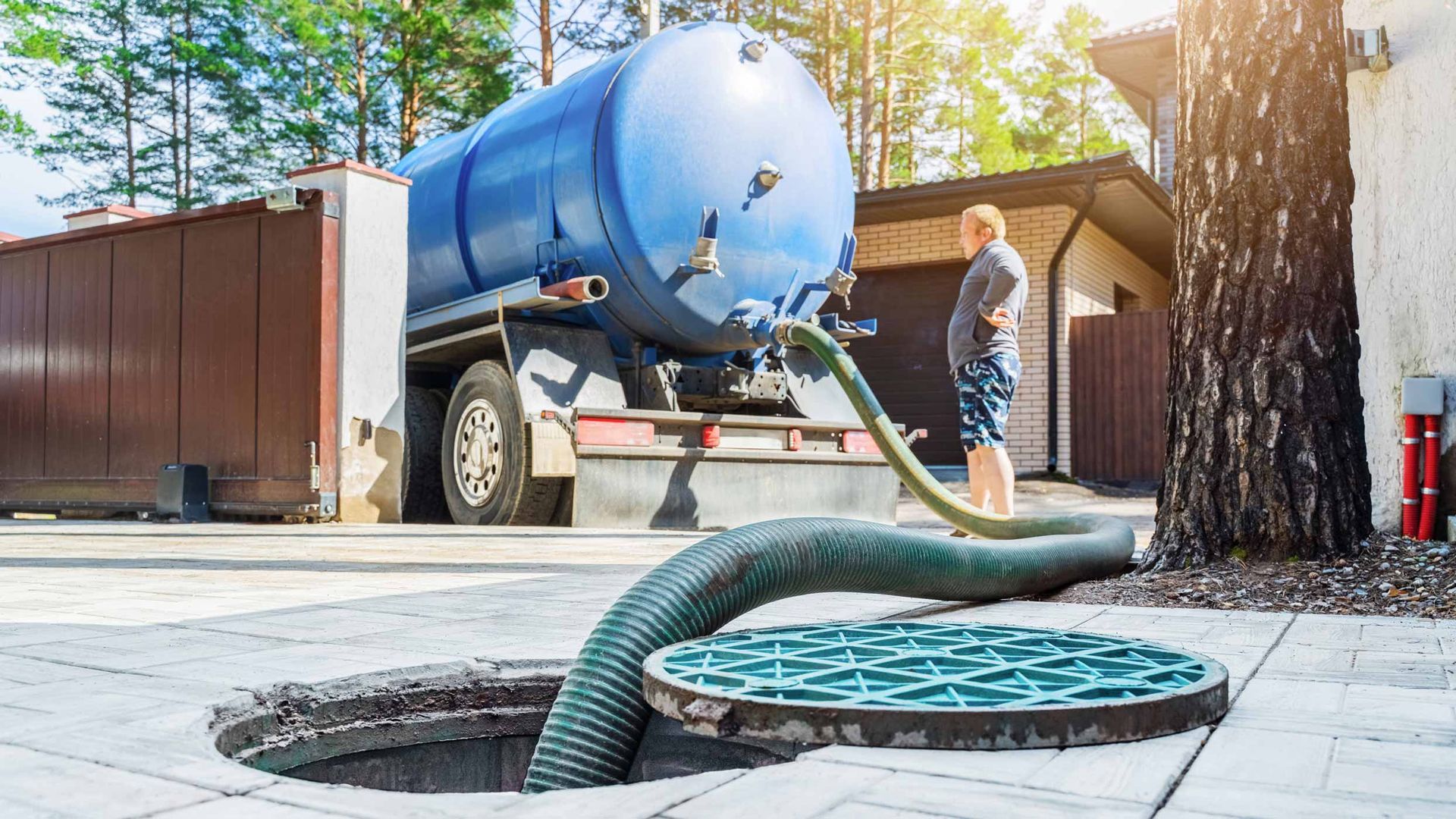 Blue vacuum truck pumping from a manhole in a driveway, with a worker nearby.