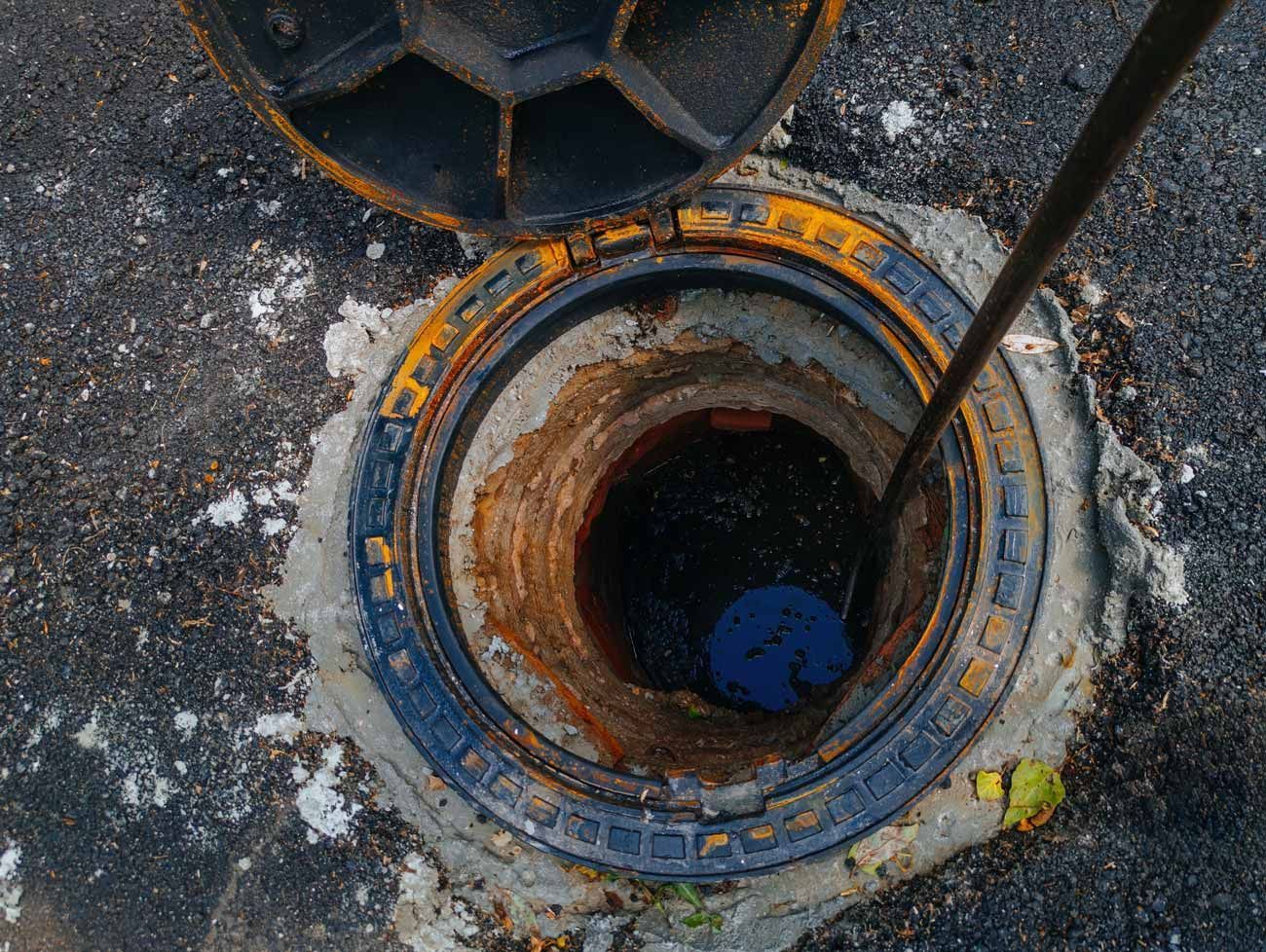 Open manhole with rusted metal rim and a blue object inside, surrounded by rough pavement.