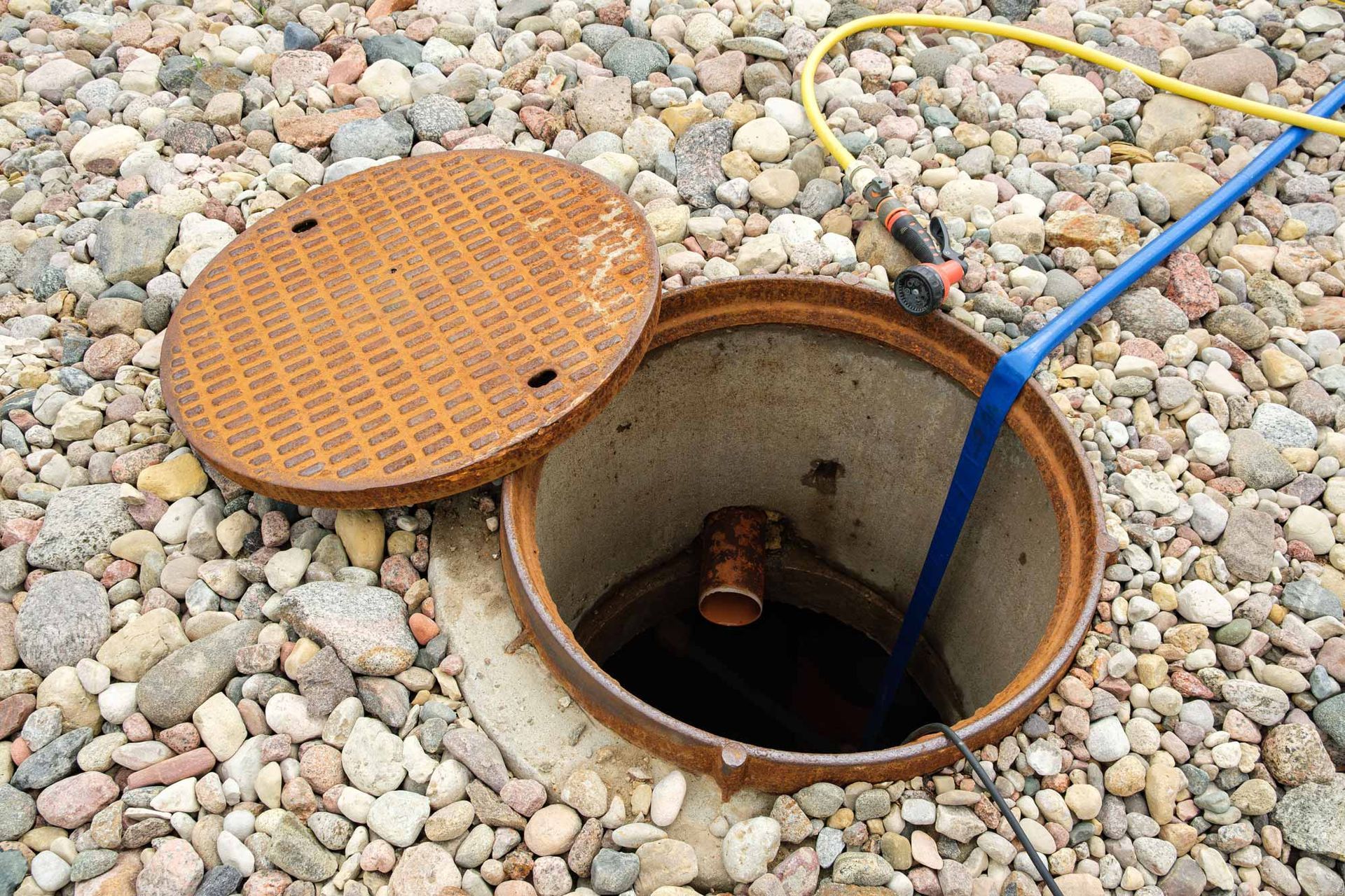 Open utility access hole with orange cover on pebble ground, blue hose inserted