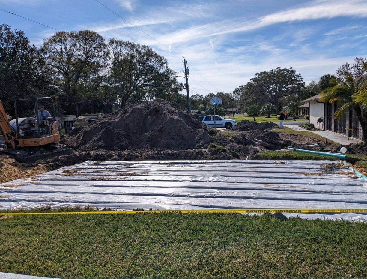 Construction site with excavator, dirt piles, and white-covered ground beside a house and trees