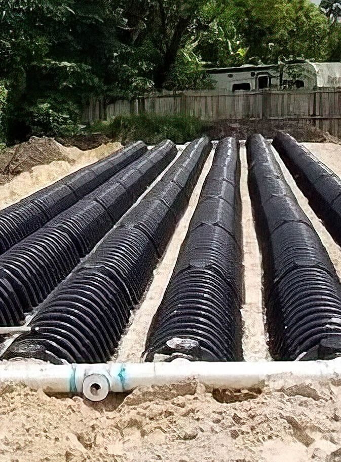 Rows of black corrugated drainage pipes laid in a sandy trench outdoors.