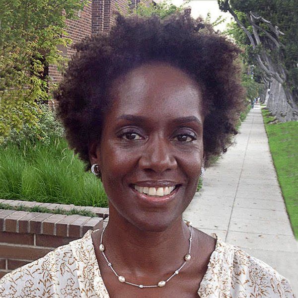A woman wearing a necklace and earrings is smiling while standing on a sidewalk.