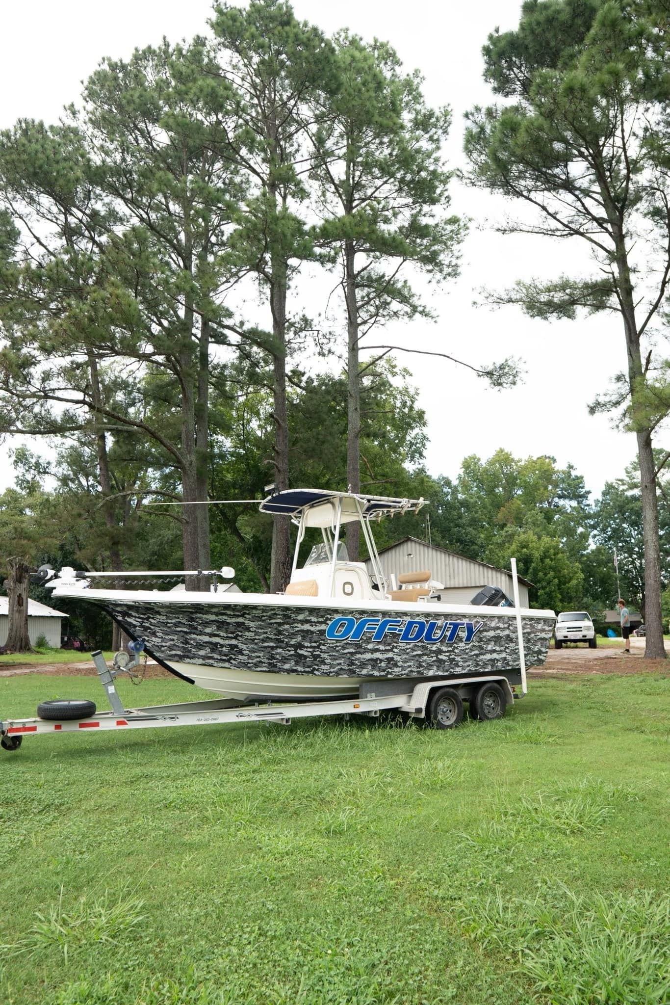 Boat on a trailer, camouflage pattern, parked on grass, trees in the background.