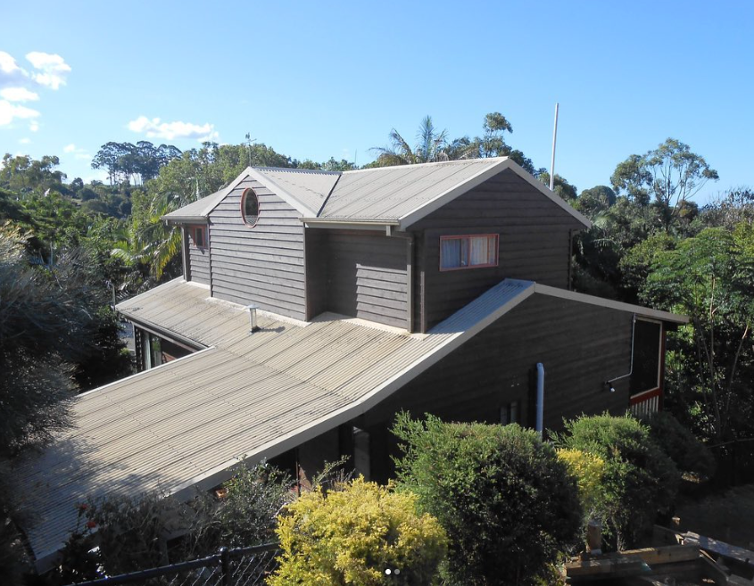 Two-story house with gray siding and corrugated roof, surrounded by green trees and blue sky.