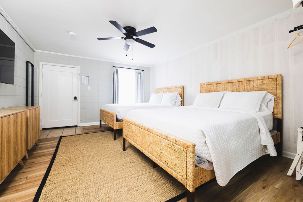 A spacious guest room at Seabirds Motel at Kure Beach featuring two queen-sized beds with rattan frames, a ceiling fan, and light wood furnishings. The room is designed for comfort and relaxation, making it ideal for a pet-friendly stay in North Carolina.