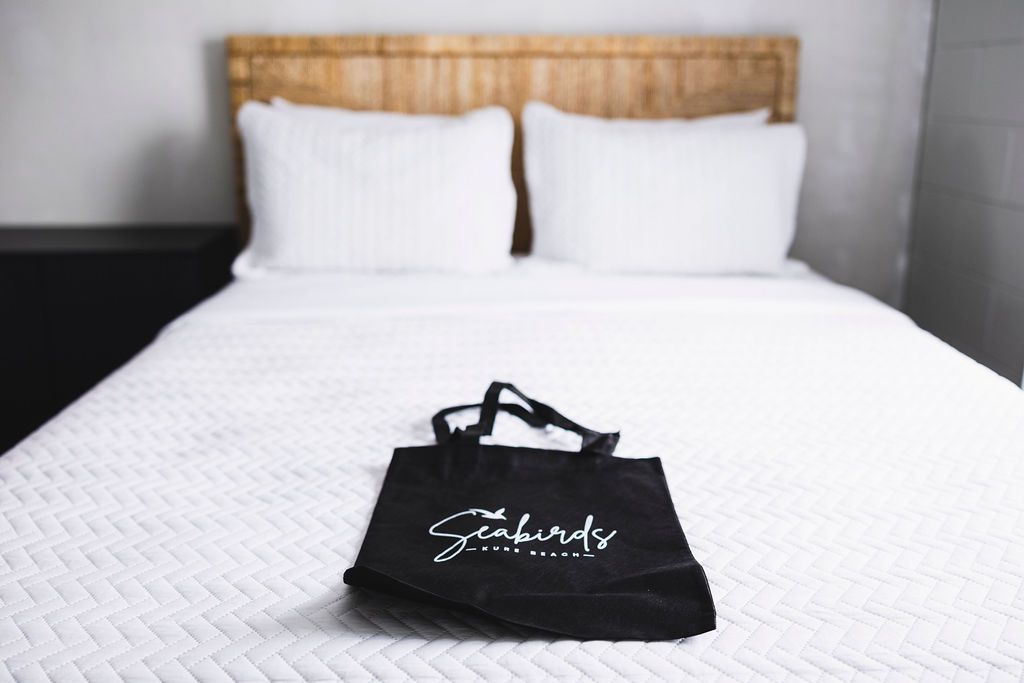 A queen-sized bed with crisp white linens at Seabirds Motel at Kure Beach with a branded tote bag on the bed. This room provides a comfortable and welcoming setting for a pet-friendly stay in North Carolina.