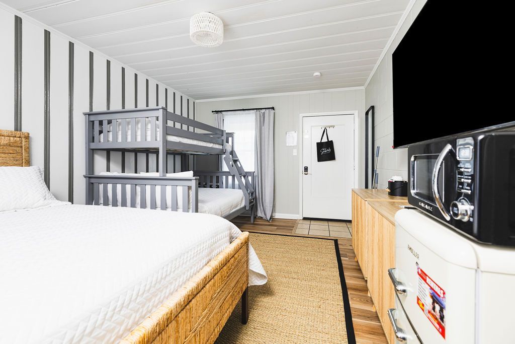 A versatile family room at Seabirds Motel at Kure Beach, showcasing a queen bed with a rattan frame, a bunk bed for additional guests, and modern amenities including a flat-screen TV and a mini-fridge. The room’s design caters to both families and pet-friendly stays in North Carolina.