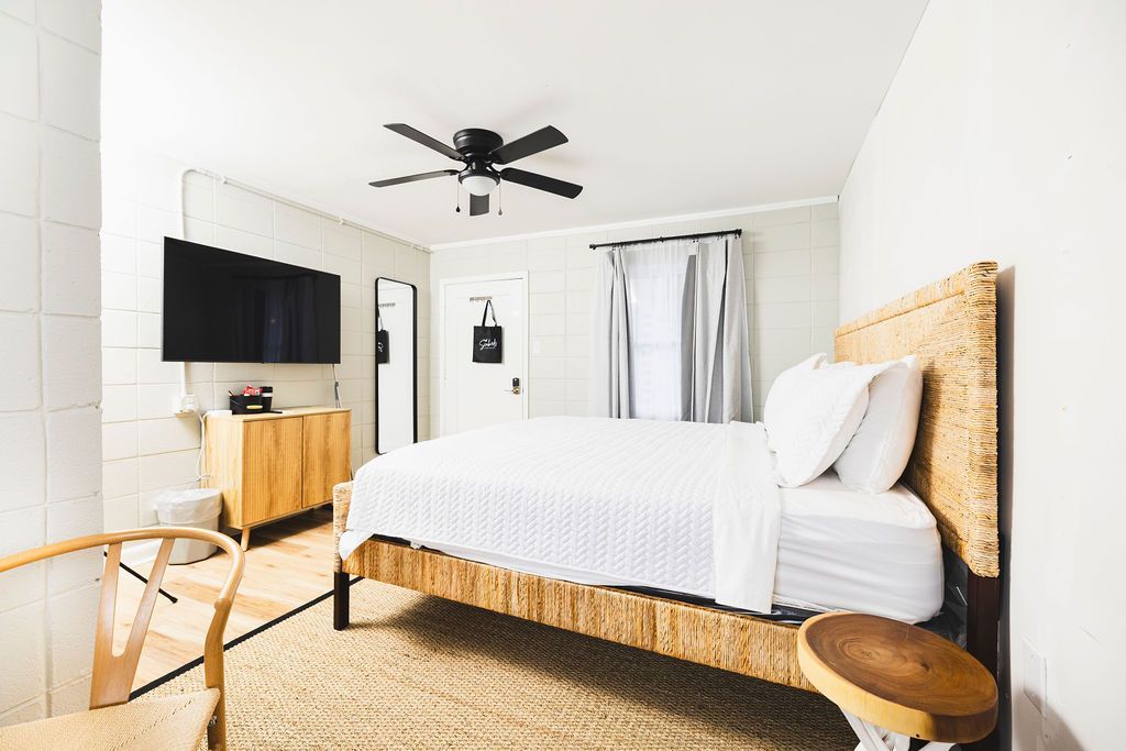 A stylish guest room at Seabirds Motel at Kure Beach with a queen-sized bed featuring a woven rattan headboard, white linens, and a mounted flat-screen TV. The room offers a modern, comfortable environment for a pet-friendly stay in North Carolina.