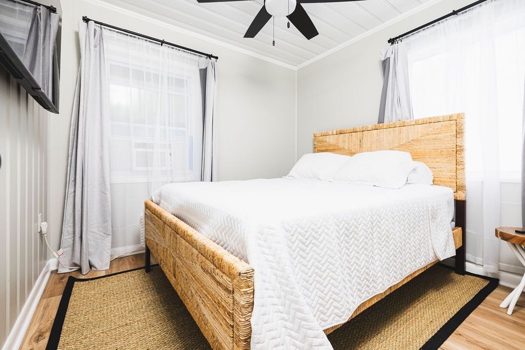 A well-appointed guest room at Seabirds Motel at Kure Beach featuring a queen-sized bed with a rattan headboard, white linens, and light-filled windows. The room provides a serene setting for a comfortable and pet-friendly lodging experience in North Carolina.