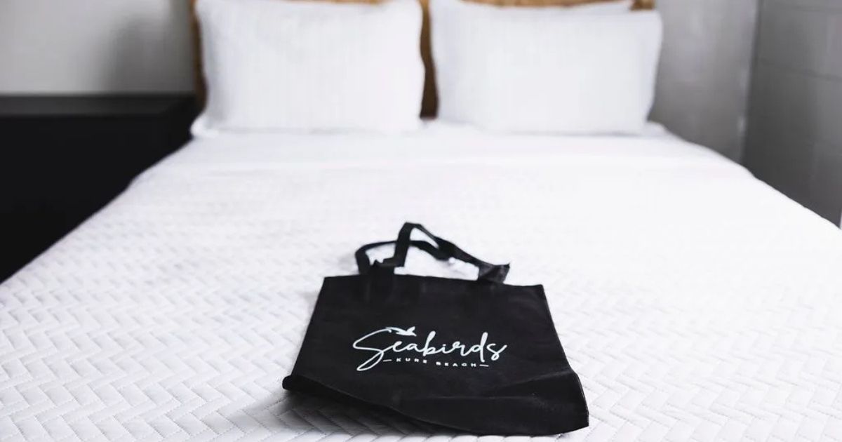 A cozy white bed at Seabirds Motel in Kure Beach, North Carolina, with a black tote bag featuring the Seabirds logo resting on the quilted bedspread.