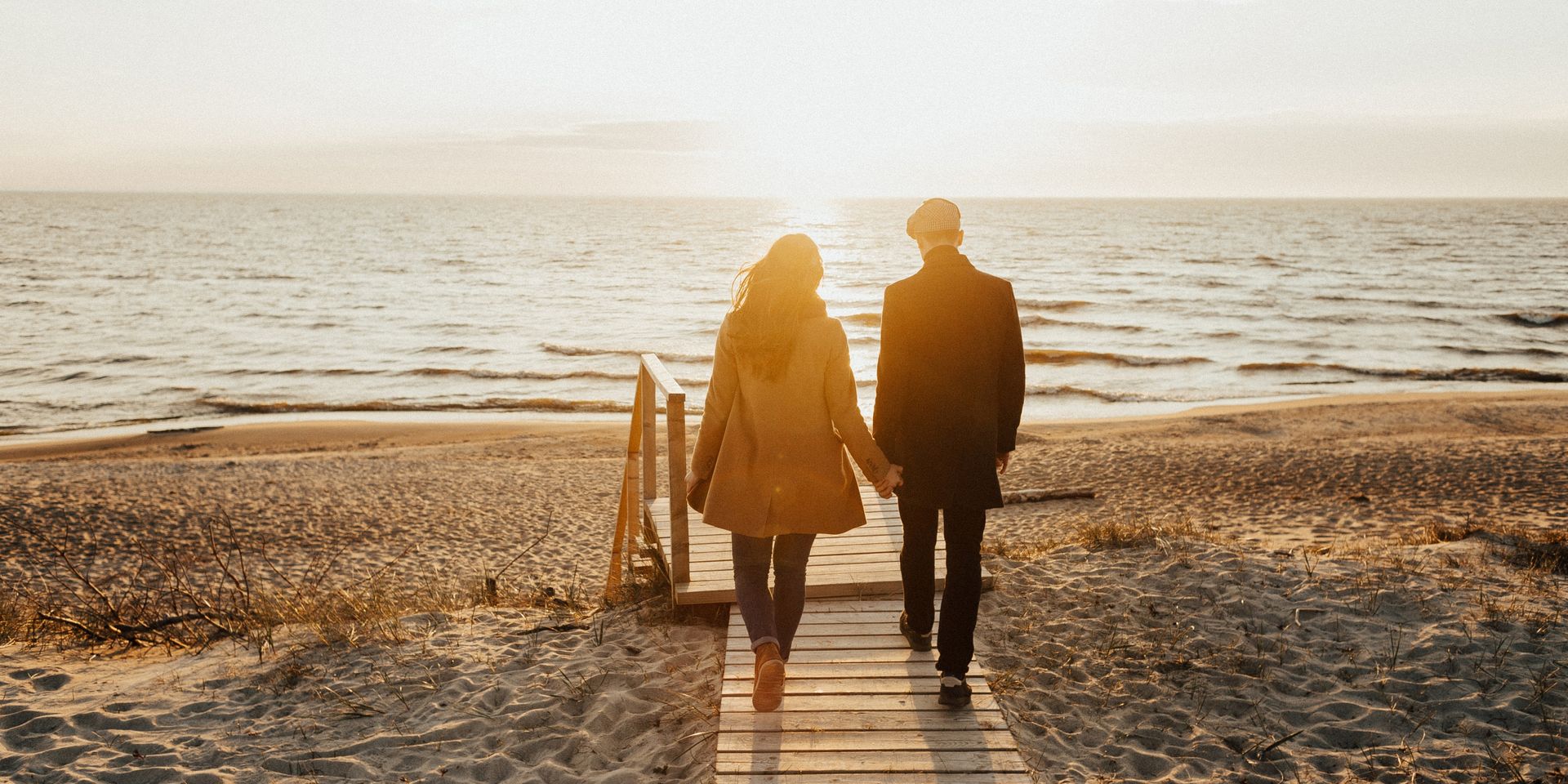 A couple walking along a wooden boardwalk leading to the winter beach bathed in the sunrise.